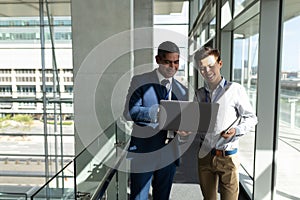 Front view of two businessman standing on the first floor walkway and working on a laptop in office