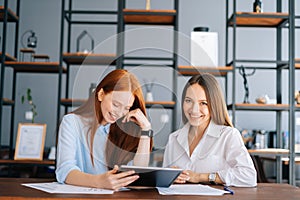 Front view of two cheerful young businesswomen working using digital tablet at meeting desk with job documents at office