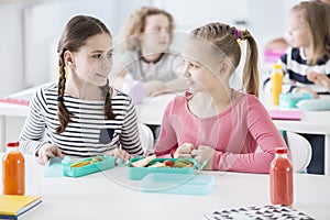 Front view of two girls sitting by a school desk with opened lunch boxes with healthy vegetables and sandwiches. Bottles