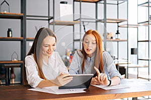 Front view of two happy young business women working using digital tablet during meeting at desk with job documents at