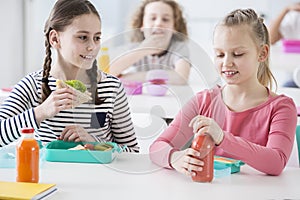 Front view of two junior girls in a school cafeteria during lunch break. One holding a wholewheat sandwich, the other opening a b