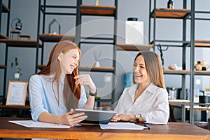 Front view of two laughing young businesswomen working using digital tablet at meeting desk with job documents at office