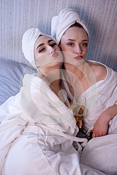 Front view of two smiling female friends with towels and in bathrobes sitting on bed and doing pedicure. Cute happy young girls