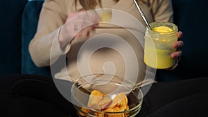 Front view unrecognizable pregnant young Caucasian woman eating honey and potato chips sitting indoors. Expectant with
