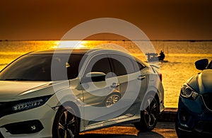 Front view of white and blue compact car parked on concrete road by sea beach at sunset sky. Electric and hybrid car technology.