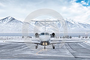 Front view of the white business jet taxiing on airport taxiway in winter on the background of high scenic snow capped mountains