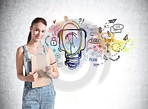 Front view of woman in denim jumpsuit holding folder smiling and standing nearby concrete wall with colorful light bulb sketch