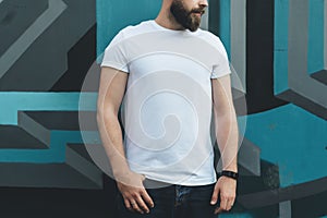 Front view. Young bearded hipster man dressed in white t-shirt is stands against wall with graffiti. Mock up.