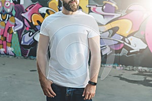 Front view. Young bearded hipster man dressed in white t-shirt is stands against wall with graffiti. Mock up.