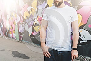Front view. Young bearded hipster man dressed in white t-shirt is stands against wall with graffiti. Mock up.