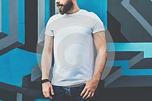 Front view. Young bearded hipster man dressed in white t-shirt is stands against wall with graffiti. Mock up.