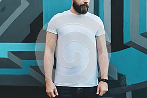 Front view. Young bearded hipster man dressed in white t-shirt is stands outdoor against wall with graffiti. Mock up.