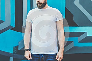 Front view. Young bearded hipster man dressed in white t-shirt is stands outdoor against wall with graffiti. Mock up.
