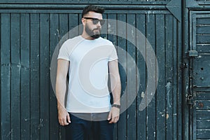 Front view. Young bearded hipster man dressed in white t-shirt and sunglasses is stands against dark wood wall.