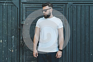 Front view. Young bearded hipster man dressed in white t-shirt and sunglasses is stands against dark wood wall.