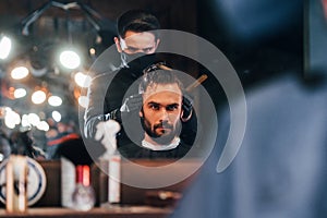 Front view of young bearded man that sitting and getting haircut in barber shop by guy in black protective mask