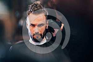 Front view of young bearded man that sitting and getting haircut in barber shop by guy in black protective mask