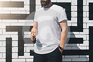 Front view. Young bearded millennial man dressed in white t-shirt is stands against brick wall. Mock up.