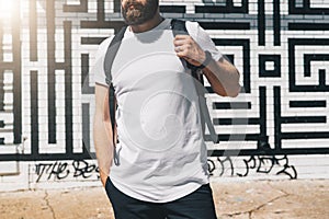 Front view. Young bearded millennial man dressed in white t-shirt is stands against brick wall. Mock up.