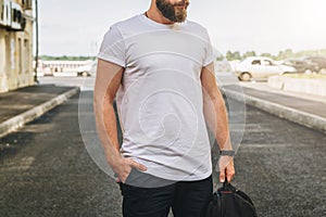 Front view. Young bearded millennial man dressed in white t-shirt is stands on city street. Mock up.