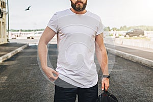 Front view. Young bearded millennial man dressed in white t-shirt is stands on city street. Mock up.