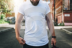 Front view. Young bearded millennial man dressed in white t-shirt is stands on city street. Mock up.