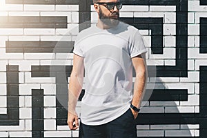 Front view. Young bearded millennial man dressed in white t-shirt and sunglasses is stands against brick wall. Mock up.