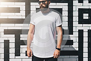 Front view. Young bearded millennial man dressed in white t-shirt and sunglasses is stands against brick wall. Mock up.