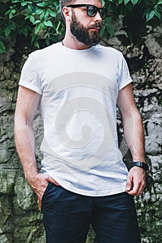 Front view. Young bearded millennial man dressed in white t-shirt and sunglasses is stands against dark wall. Mock up.