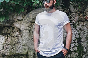 Front view. Young bearded millennial man dressed in white t-shirt and sunglasses is stands against dark wall. Mock up.
