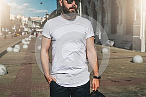 Front view. Young bearded millennial man dressed in white t-shirt and sunglasses is stands on city street. Mock up.