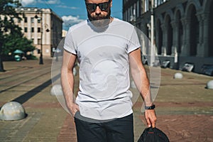 Front view. Young bearded millennial man dressed in white t-shirt and sunglasses is stands on city street. Mock up.