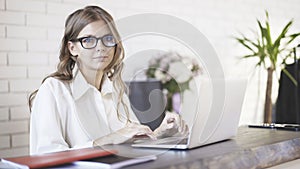 Front view of a young beautiful woman wearing glasses typing at her laptop in office and looking at camera.