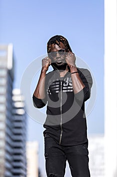 Front view of a young black african man wearing casual clothes and sunglasses standing in the street while looking camera in a