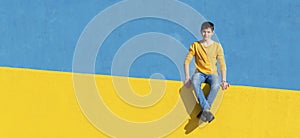 Front view of a young boy wearing casual clothes sitting on a yellow fence against a blue wall in a sunny day