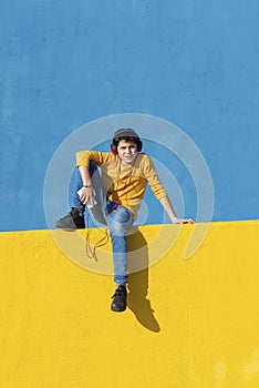 Front view of a young boy wearing casual clothes sitting on a yellow fence against a blue wall while using a mobile phone to