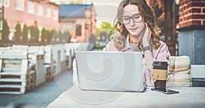 Front view.Young businesswoman with glasses is sitting at table in street cafe and uses laptop.