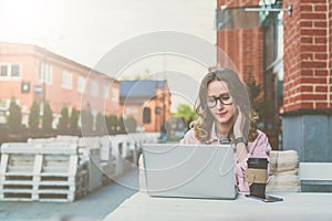 Front view.Young businesswoman with glasses is sitting at table in street cafe and uses laptop.