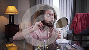 Front view of young Caucasian man looking at mirror and applying mascara on mustaches. Intersex person doing makeup on