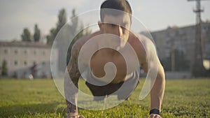 Front view of young concentrated man pushing up on green field outdoors. Portrait of confident Caucasian sportsman