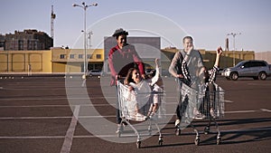 Front view of a young friends having fun outdoors on shopping trolleys. Multiethnic young people racing on shopping