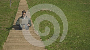 Front View of Young Hipster Man In Sunglasses Sitting On The Stairs Using Laptop