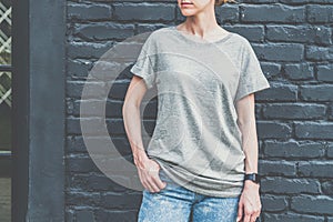 Front view. Young millennial woman dressed in gray t-shirt is stands against dark brick wall.