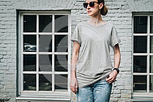 Front view. Young millennial woman dressed in gray t-shirt is stands against gray brick wall.