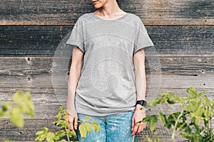 Front view. Young millennial woman dressed in gray t-shirt is stands against gray wood wall.