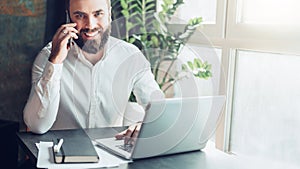 Front view. Young smiling bearded businessman is sitting in office at table in front of laptop, talking on cell phone.