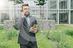 Young smiling businessman in suit and tie is standing outdoor,holding tablet computer and talking on his cell phone