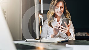 Front view. Young smiling businesswoman is sitting in office at table in front of computer, using smartphone.