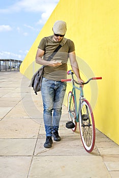 Front view of a young trendy man with a fixed bike wearing casual clothes while using a mobile phone against a yellow wall