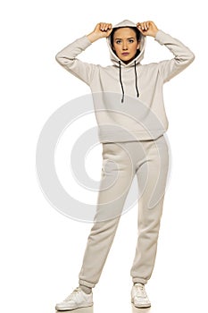 Front view of a young woman in a gray tracksuit and hood posing to a white background in the studio